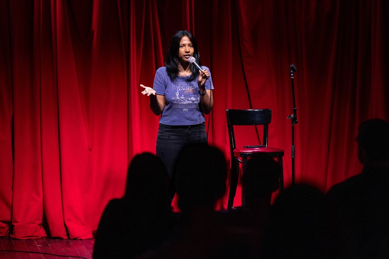 A comic on stage at the first Baklava Comedy show, red curtain behind her, the room in silhouette